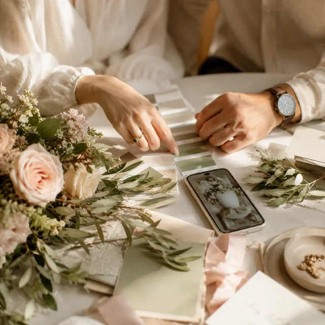 Hände wählen mit Farbmustern, Blumen und Smartphone das Design einer Hochzeitshomepage aus.