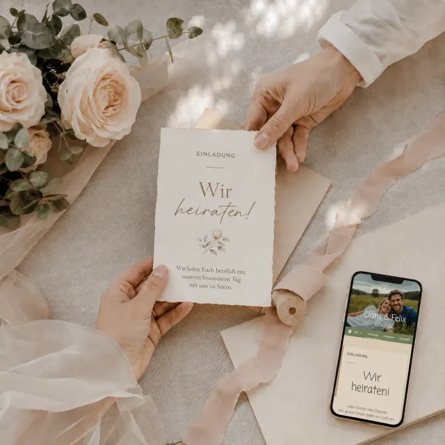Hände ordnen eine schlichte Hochzeitskarte und ein Smartphone zwischen Blumen und Seidenband
